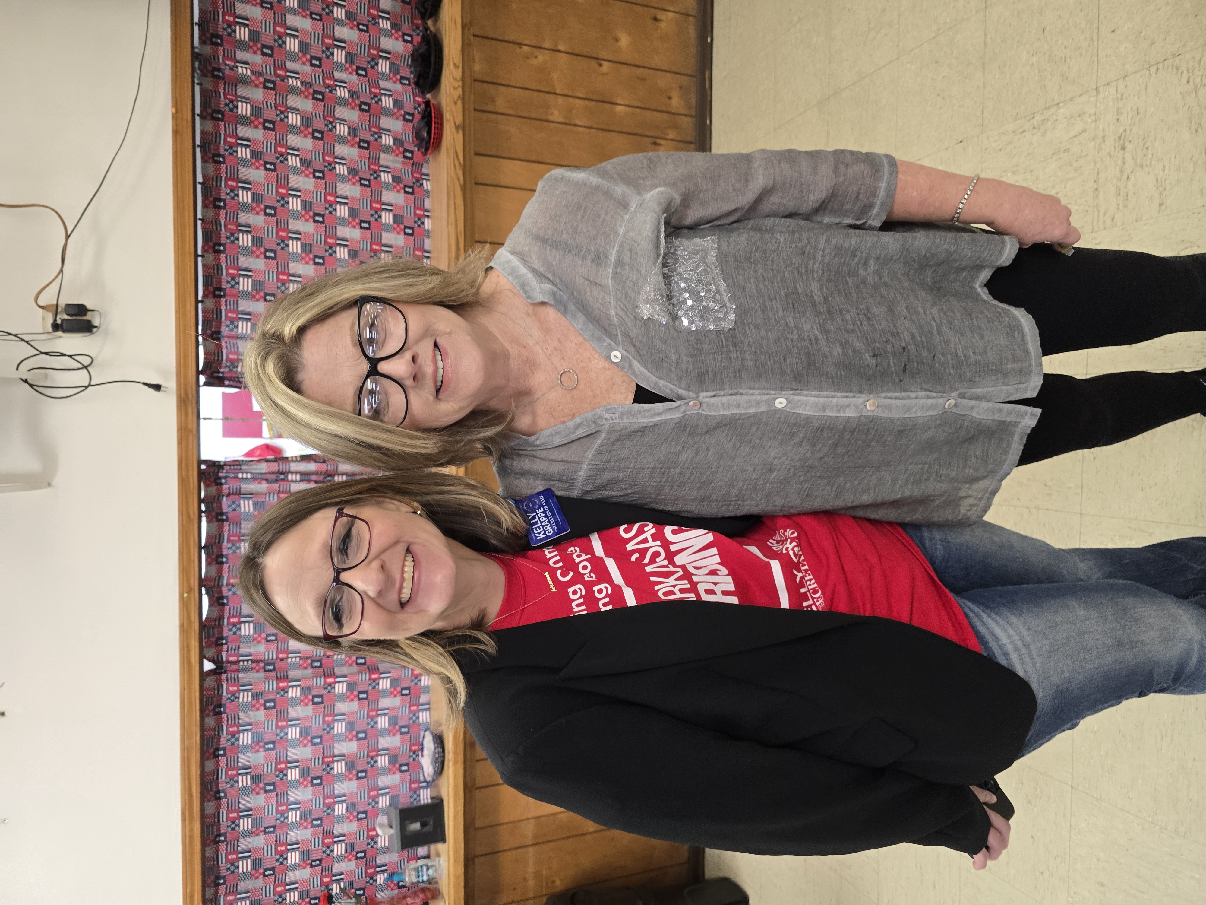 Kelly Grappe in an Arkansas Rising campaign shirt poses with a supporter inside a community hall decorated with American flags.