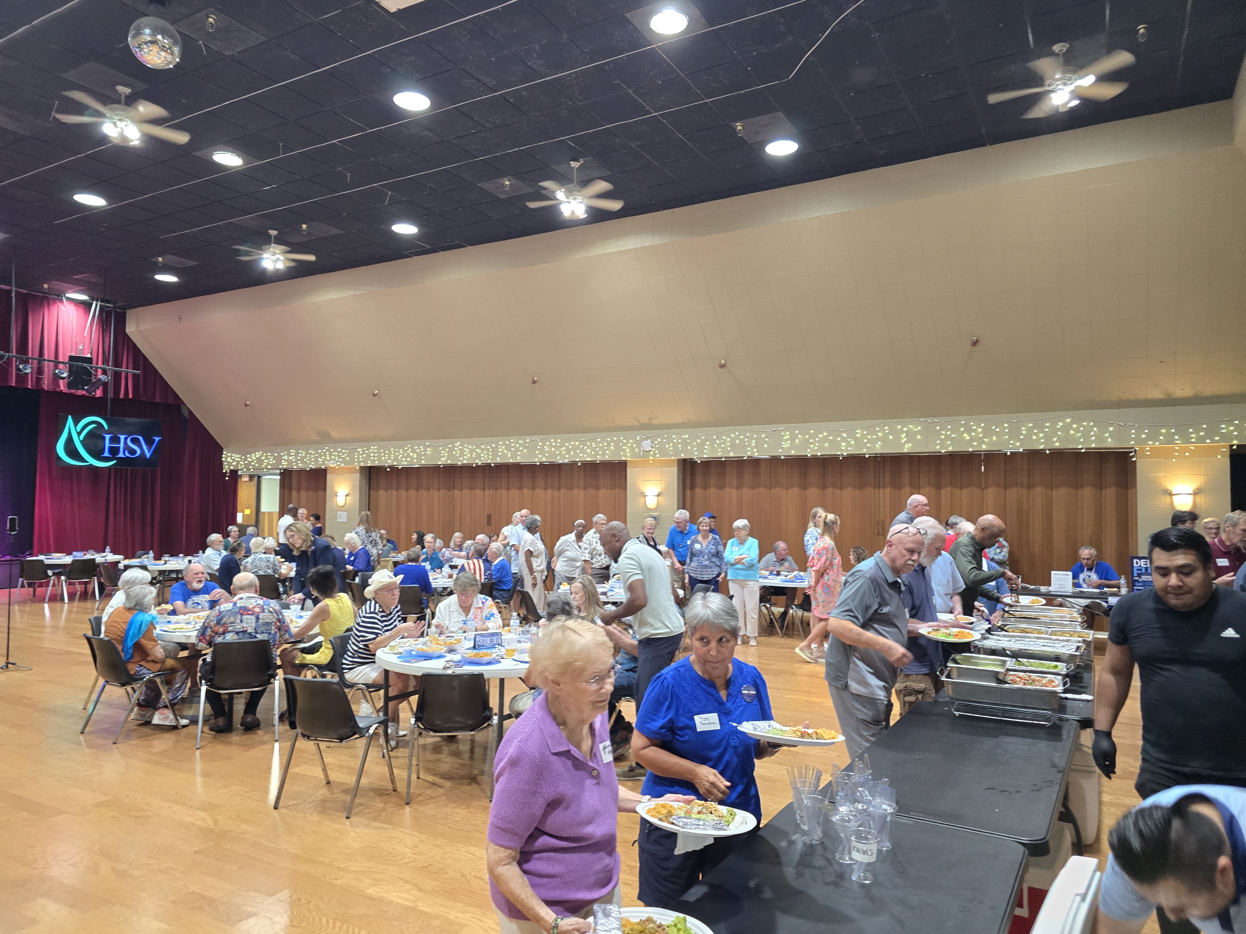 Community members line up at buffet tables and sit at round tables during a crowded Hot Springs Village dinner event.