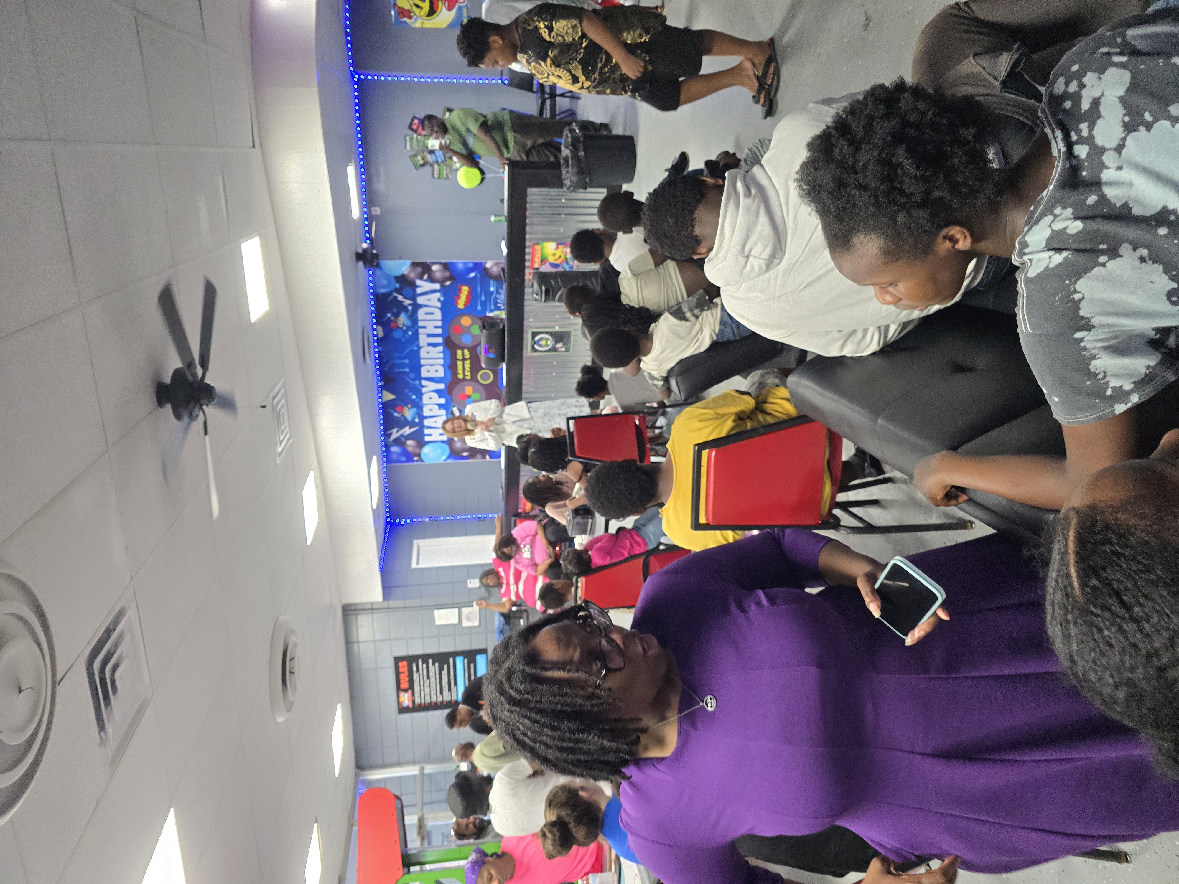 Kelly Grappe speaks into a microphone at a community center counter while families and children listen from tables.