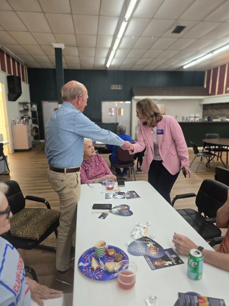 Kelly Grappe for Arkansas Secretary of State — campaign photo Kelly Grappe, candidate for Arkansas Secretary of State. Kelly Grappe shakes hands with a community member beside a hall table of campaign materials.