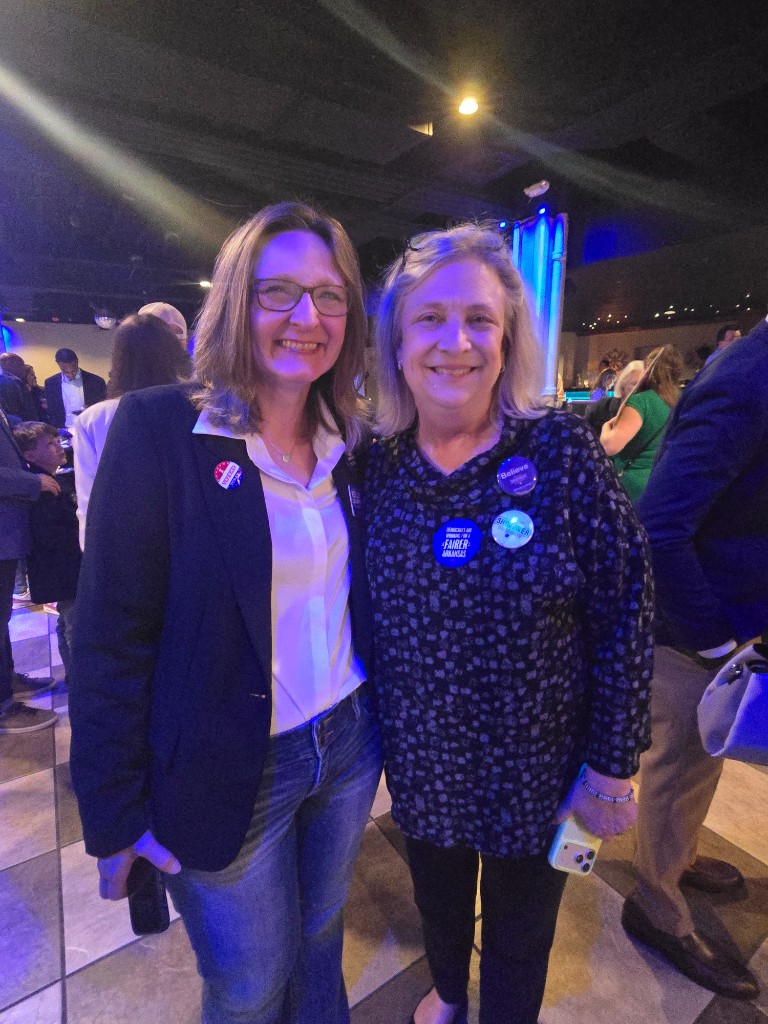 Kelly Grappe for Arkansas Secretary of State — campaign photo Kelly Grappe, candidate for Arkansas Secretary of State. Kelly Grappe smiles beside a supporter, wearing an I Voted sticker on her blazer at an indoor event.