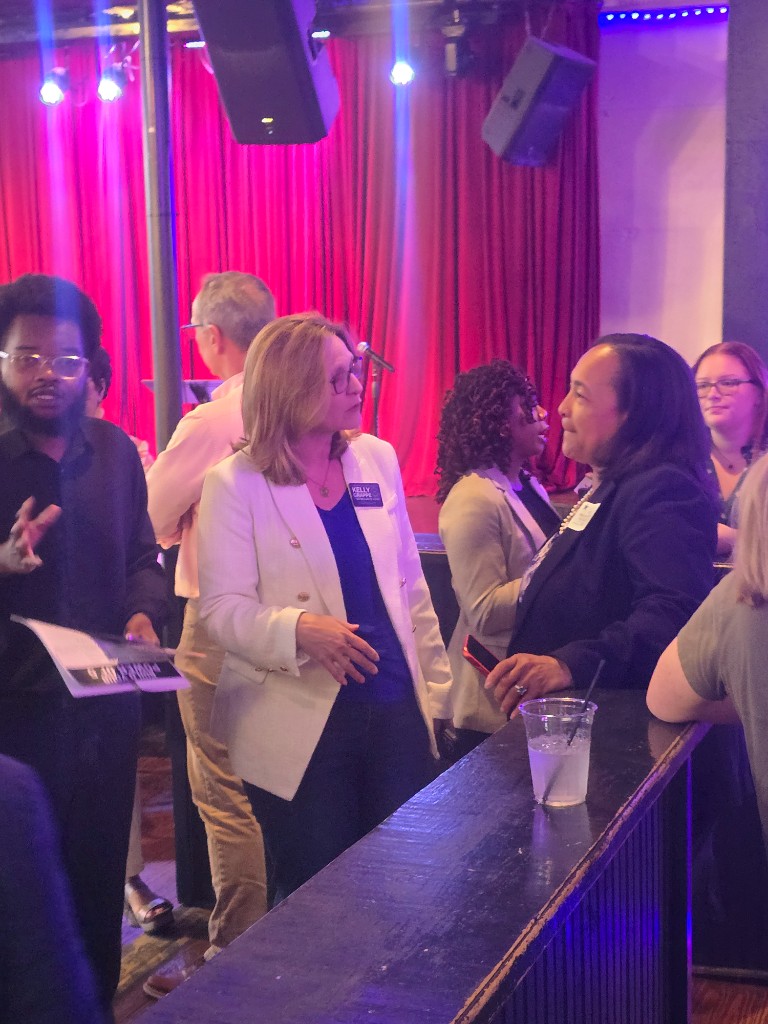 Kelly Grappe for Arkansas Secretary of State — campaign photo Kelly Grappe, candidate for Arkansas Secretary of State. Kelly Grappe in a white blazer listens to a community member near a stage at an indoor event.