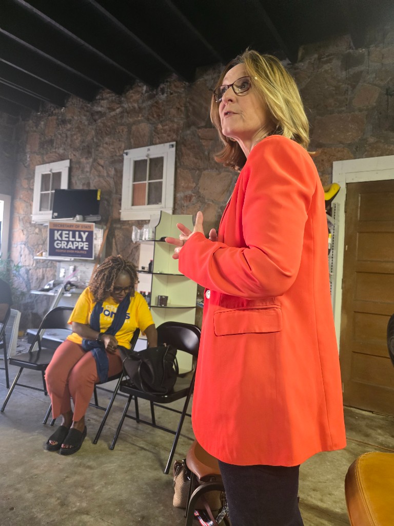 Kelly Grappe for Arkansas Secretary of State — campaign photo Kelly Grappe speaks indoors in a red-orange blazer beside her People Over Politics Secretary of State campaign sign.