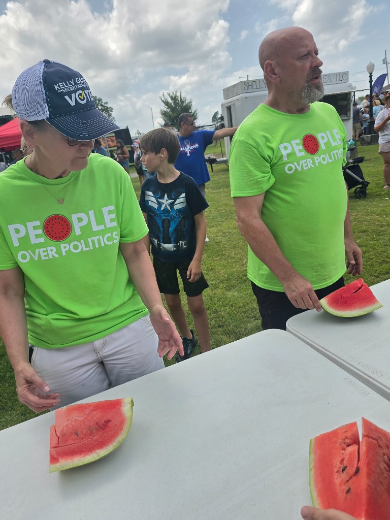 Kelly Grappe in a People Over Politics watermelon shirt serves watermelon slices at an outdoor festival table.